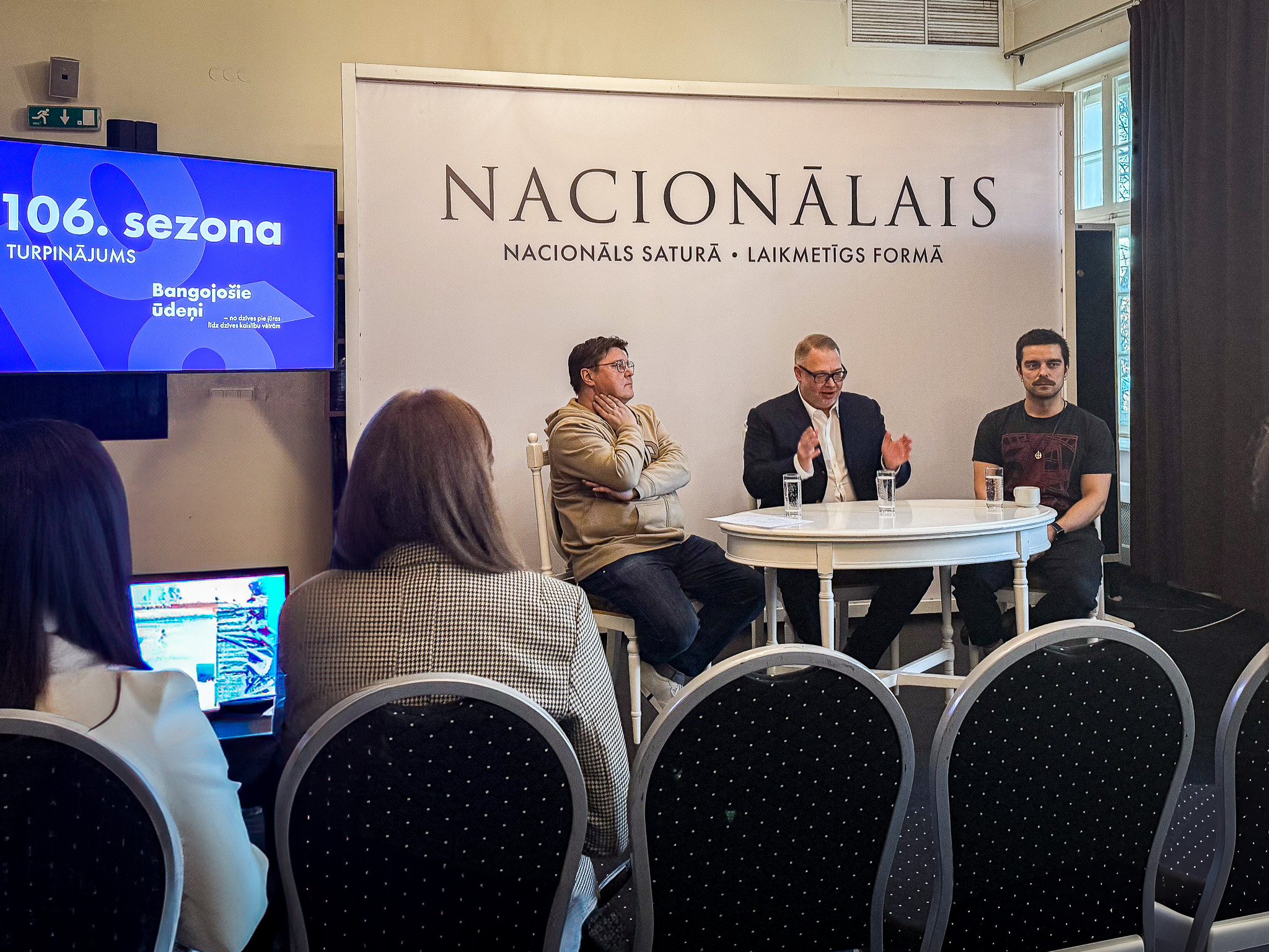 Trīs vīrieši pie apaļa galda preses konferencē, fonā uzraksts Nacionālais un ekrāns ar sezonas informāciju