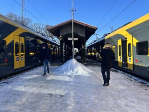 Ziemā apsnigusi dzelzceļa stacijas platforma, kur divi dzeltenmelni pasažieru vilcieni stāv pretim viens otram, un cilvēki soļo pa apledojušu peronu