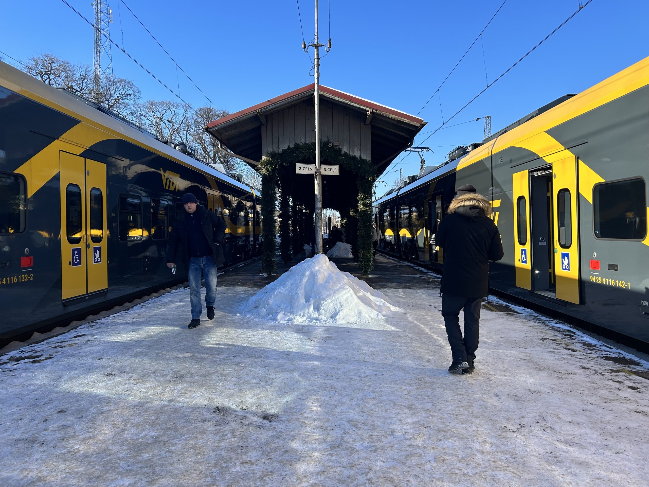 Ziemā apsnigusi dzelzceļa stacijas platforma, kur divi dzeltenmelni pasažieru vilcieni stāv pretim viens otram, un cilvēki soļo pa apledojušu peronu
