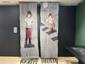 Divi vertikāli gobelēni ar cilvēku figūrām – viens attēlo stāvošu zēnu, otrs meiteni, kas skrien ar balonu.
