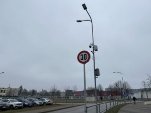 Ceļa zīme ar ātruma ierobežojumu 30 km/h, uzstādīta uz staba ar novērošanas kamerām pilsētvidē.