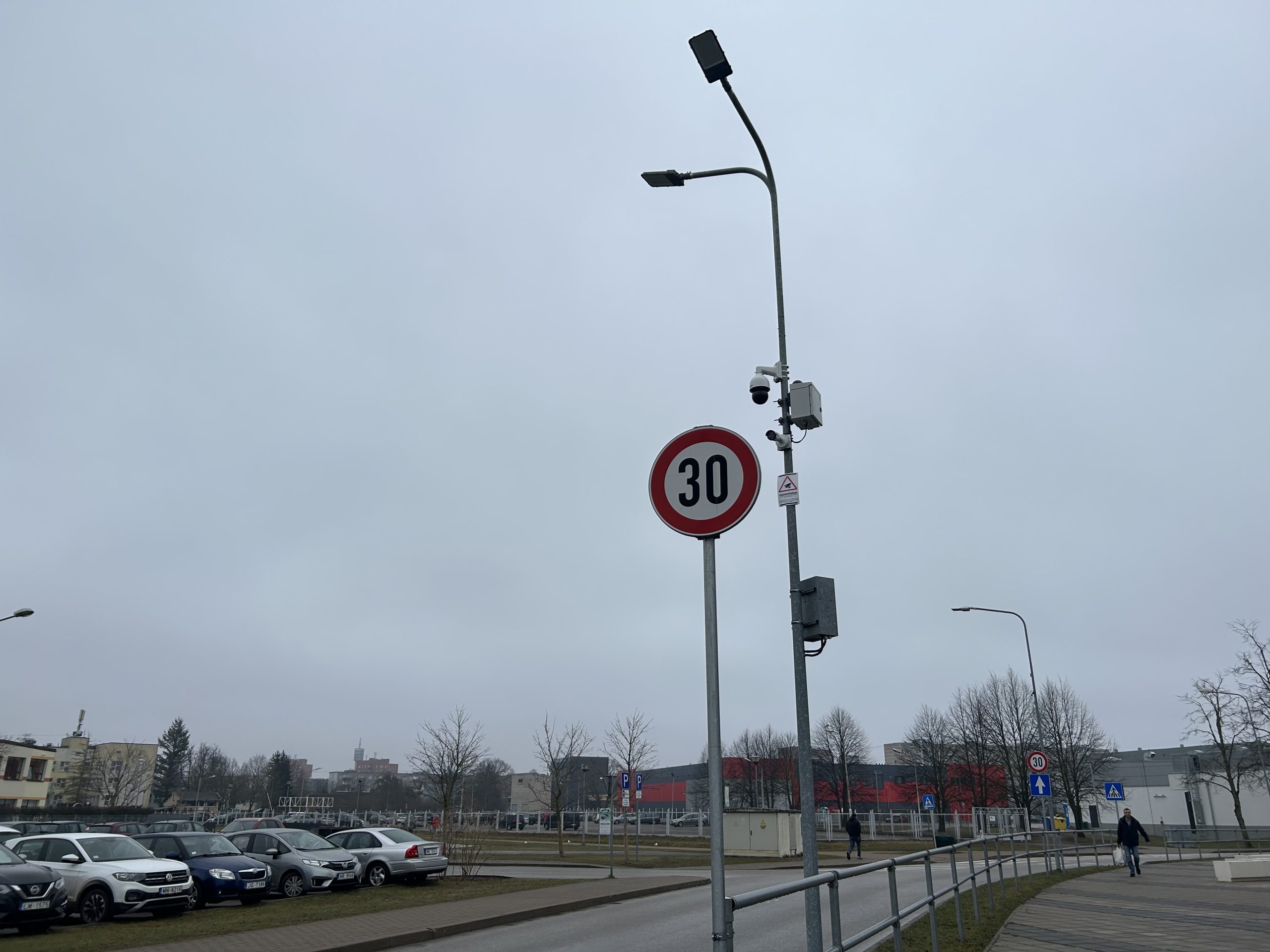 Ceļa zīme ar ātruma ierobežojumu 30 km/h, uzstādīta uz staba ar novērošanas kamerām pilsētvidē.