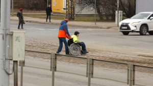 Cilvēks palīdz ratiņkrēslā esošai personai šķērsot ielu.