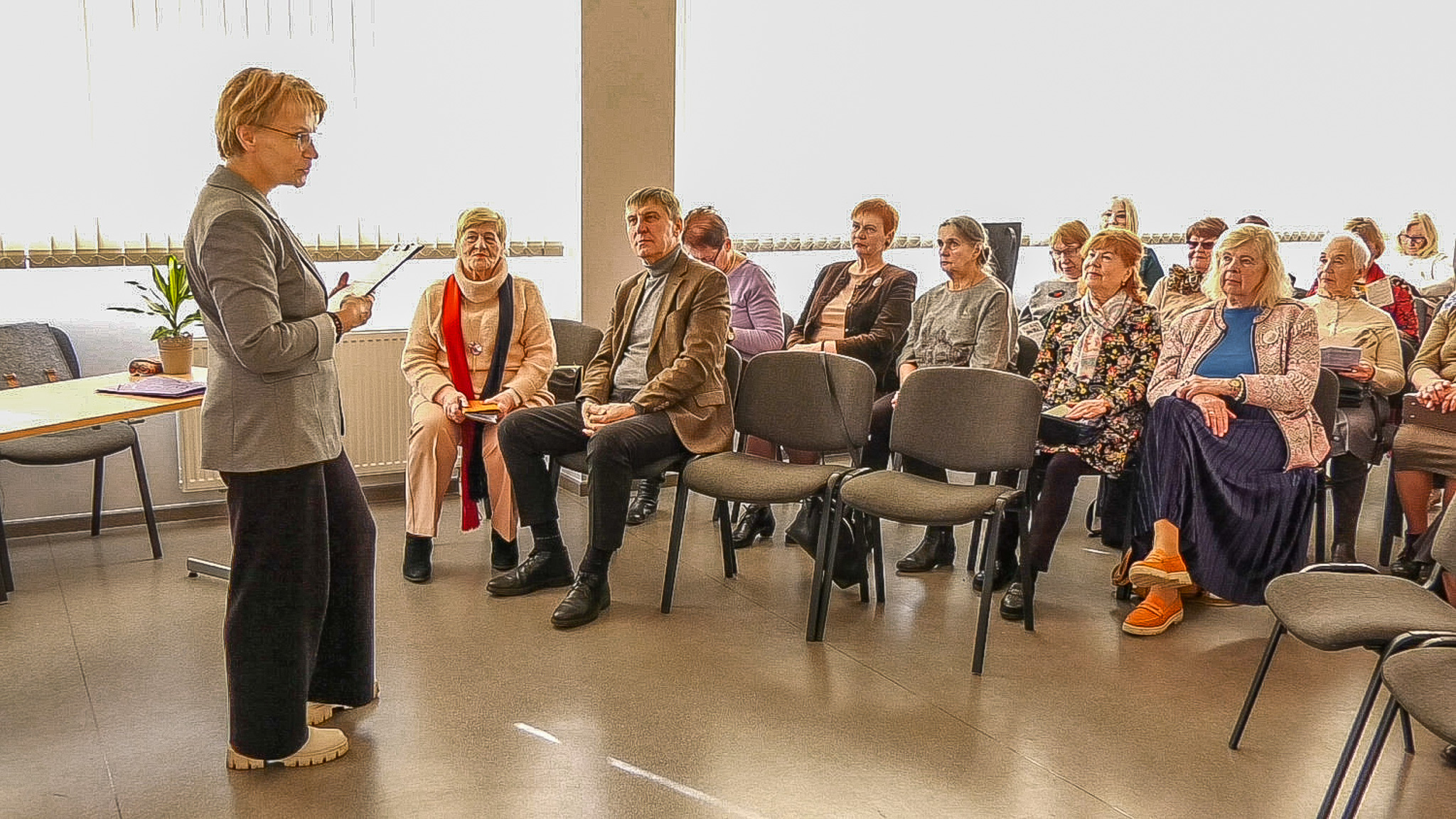 Sieviete vada sarunu vai lekciju, auditorijā sēž galvenokārt vecāka gadagājuma cilvēki