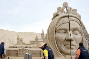 "Vairāki mākslinieki veido monumentālu smilšu skulptūru: sievietes galva ar aizvērtām acīm un uz tās izvietotas pilsētas ēku silueti."