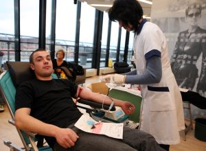 "Jauns vīrietis, atgūlies ziedošanas krēslā, ziedo asinis, kamēr medicīnas darbiniece ar cimdiem rūpējas par adatas ievietošanas vietu