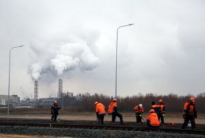 Dzelzceļa strādnieki spilgti oranžās vestēs strādā uz sliedēm rūpnieciskā vidē ar dūmojošām rūpnīcas caurulēm fonā
