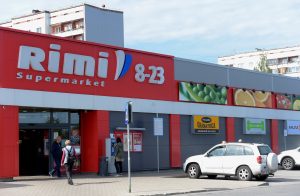 Rimi Supermarket sarkana fasāde ar uzrakstu “8–23”, pie ieejas durvīm klienti dodas iepirkties, priekšplānā automašīna