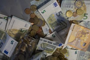 Naudas uzkrājumi stikla burkā un uz galda izkaisītas eiro banknotes un monētas