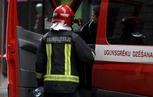Ugunsdzēsējs ar sarkanu ķiveri un aizsargtērpu stāv pie ugunsdzēsēju auto durvīm, uz kura redzams uzraksts “Ugunsgrēku dzēšana”