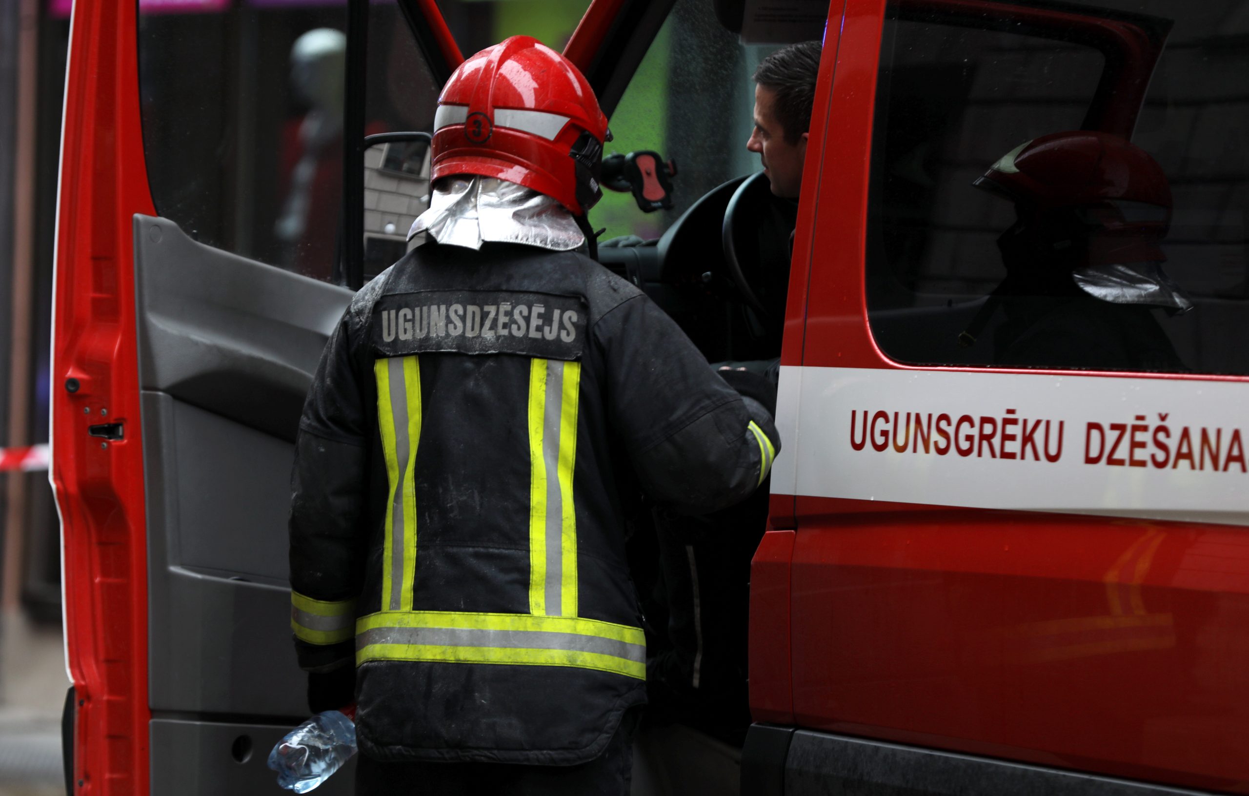 Ugunsdzēsējs ar sarkanu ķiveri un aizsargtērpu stāv pie ugunsdzēsēju auto durvīm, uz kura redzams uzraksts “Ugunsgrēku dzēšana”