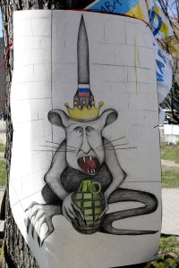 Karikatūra ar žurku cilvēka sejā, kroni un granātu, piestiprināta pie koka.