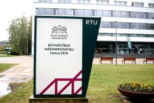 Rīgas Tehniskās universitātes Būvniecības inženierzinātņu fakultātes ēka ar informatīvo stendu priekšplānā.