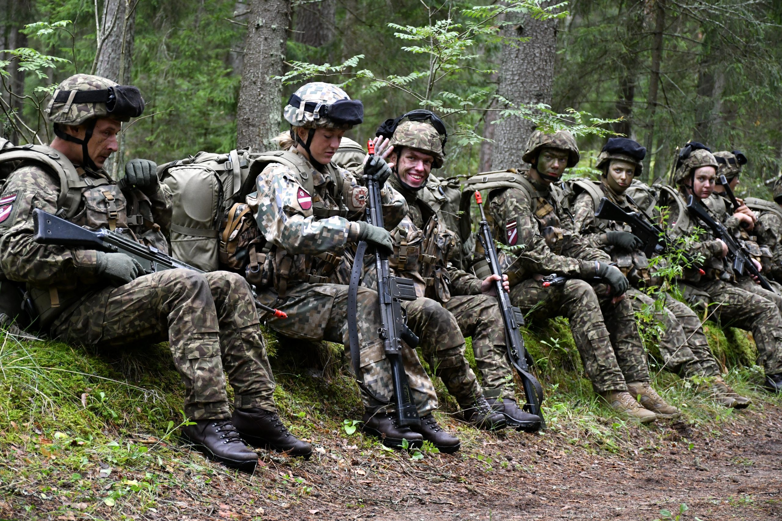 Latvijas bruņoto spēku karavīri, tērpušies kamuflāžas formās un bruņoti ar automātiskajiem ieročiem, sēž mežā lauka mācību pārtraukumā