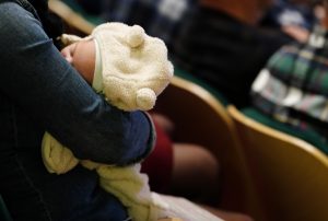 Māmiņa tur zīdaini rokās publiskā pasākumā.
