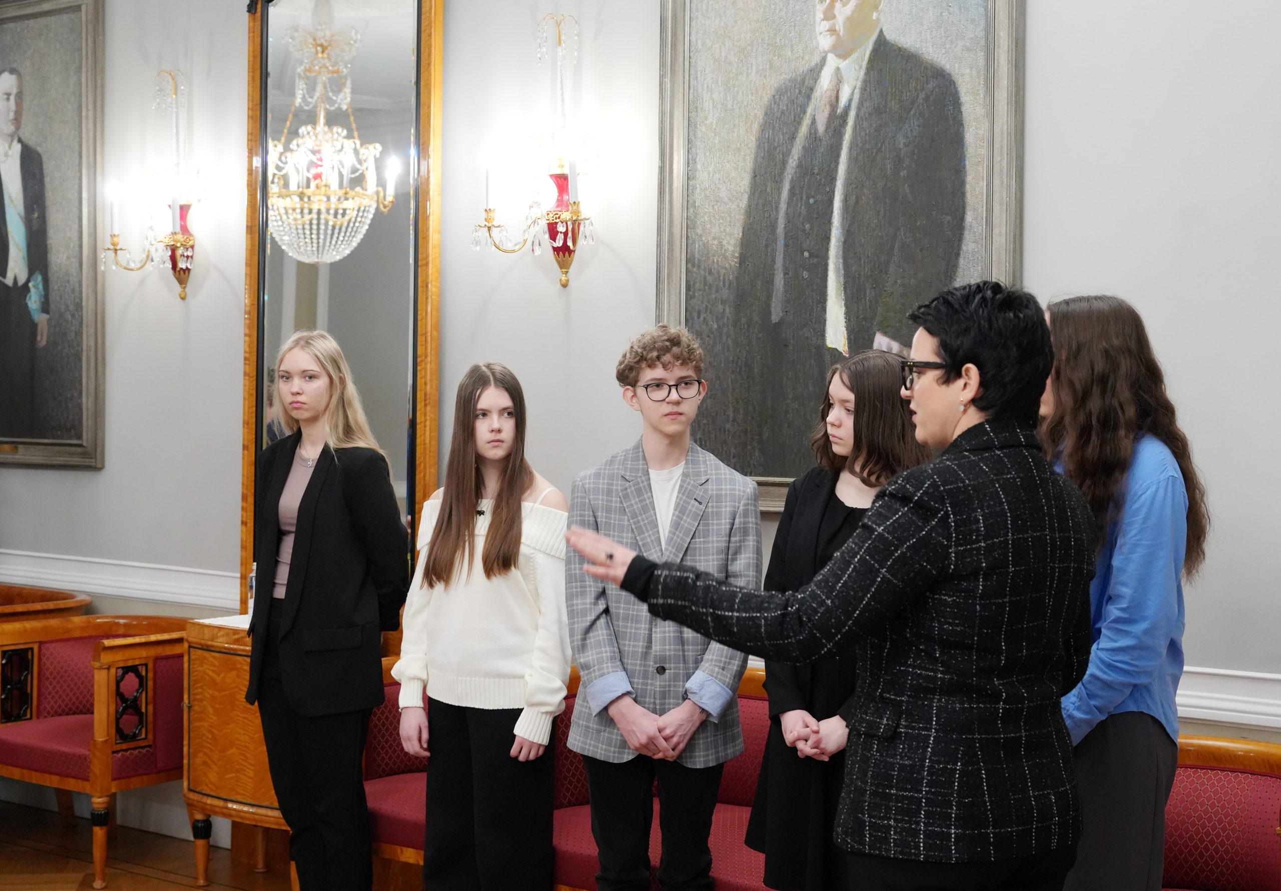 Grupa pusaudžu oficiālā telpā ar lieliem portretiem fonā, klausoties pieauguša pārstāvja skaidrojumā.