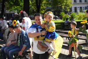 Sieviete tur mazu meiteni Ukrainas tautastērpā, apkārt cilvēki ar saulespuķēm