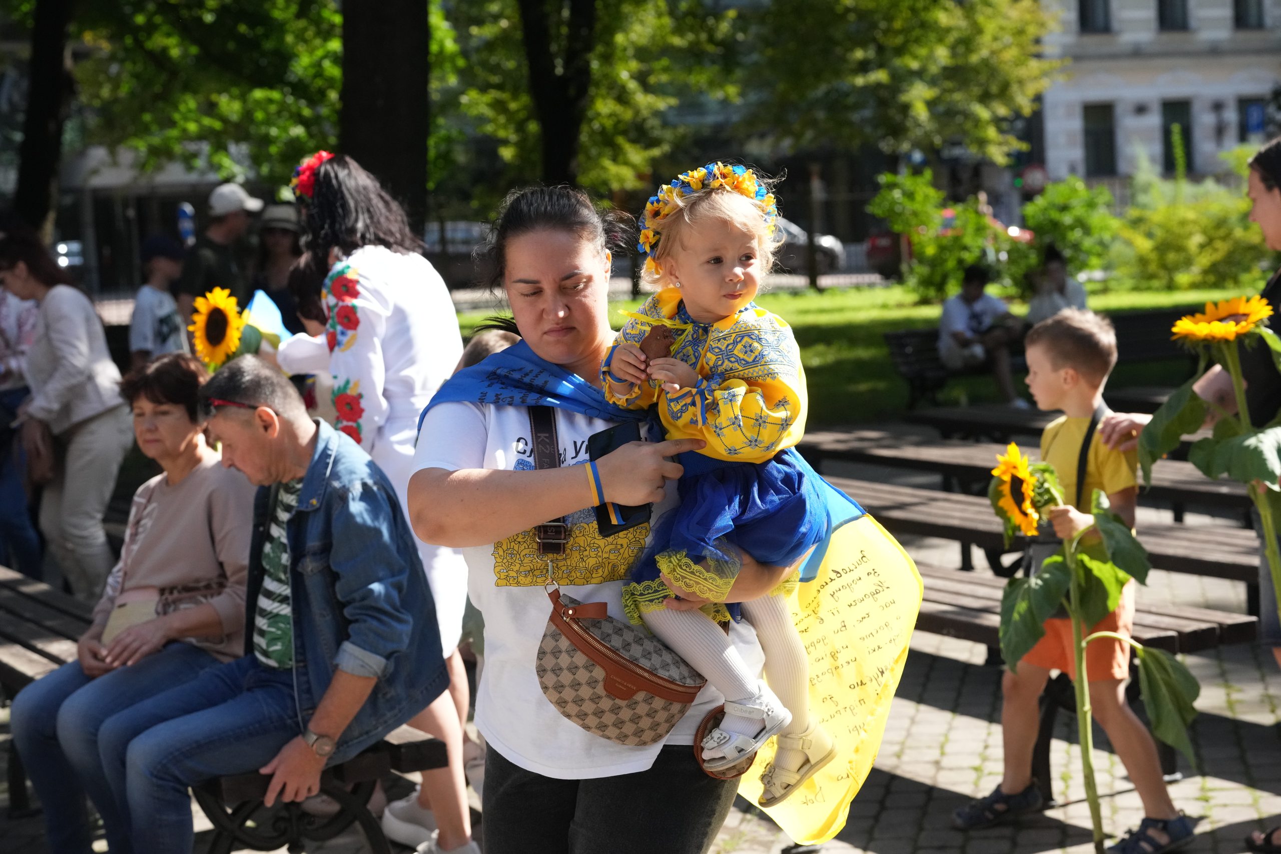 Sieviete tur mazu meiteni Ukrainas tautastērpā, apkārt cilvēki ar saulespuķēm