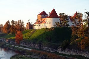 "Vēsturisks pils komplekss ar sarkanu dakstiņu jumtu un tornīšiem, kas rudenī atrodas uz zaļa kalna virs upes, apjozts ar krāsainiem kokiem."