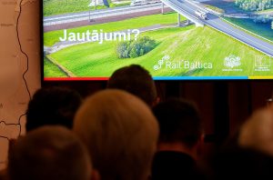 Cilvēku auditorija klausās "Rail Baltica" prezentāciju ar jautājumu slaidu.