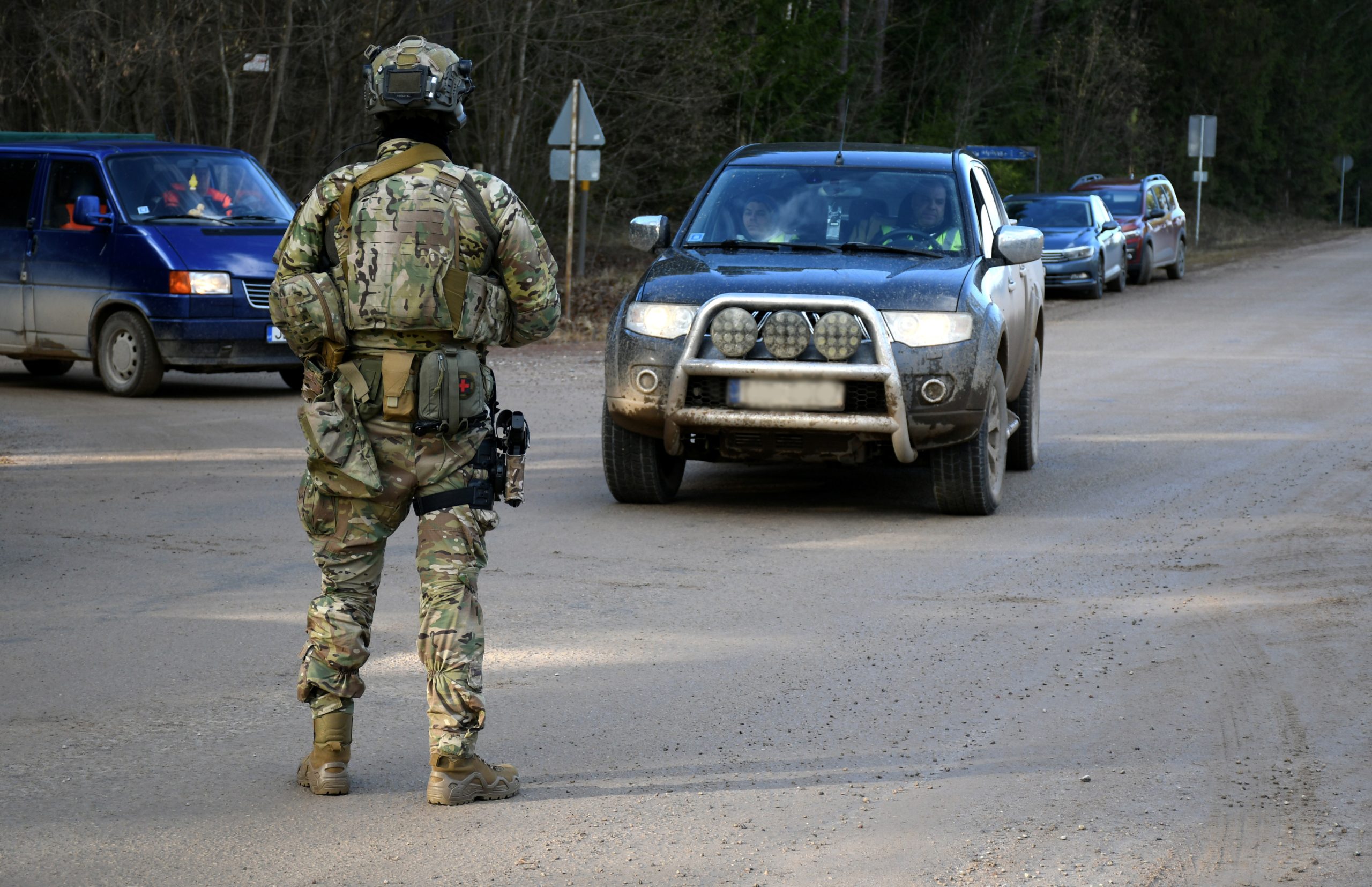 Kamuflāžas tērpā ģērbts karavīrs ar ekipējumu stāv uz lauku ceļa, kur vairākas automašīnas gaida drošības pārbaudi.
