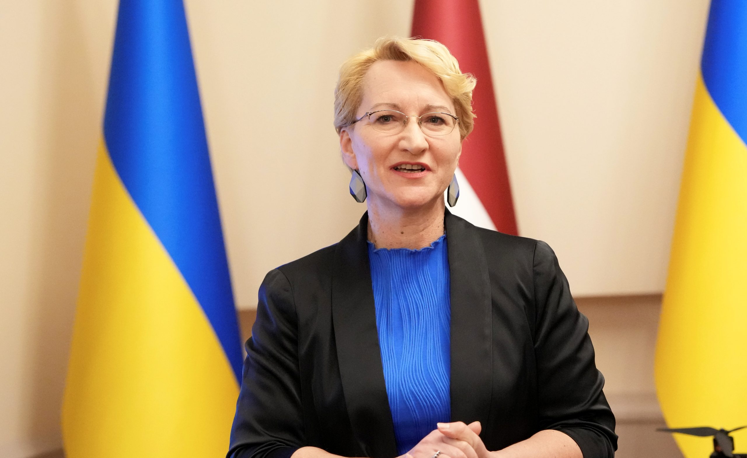 Latvijas izglītības un zinātnes ministre melnā žaketē un zilā blūzē runā, fonā Latvijas un Ukrainas karogi