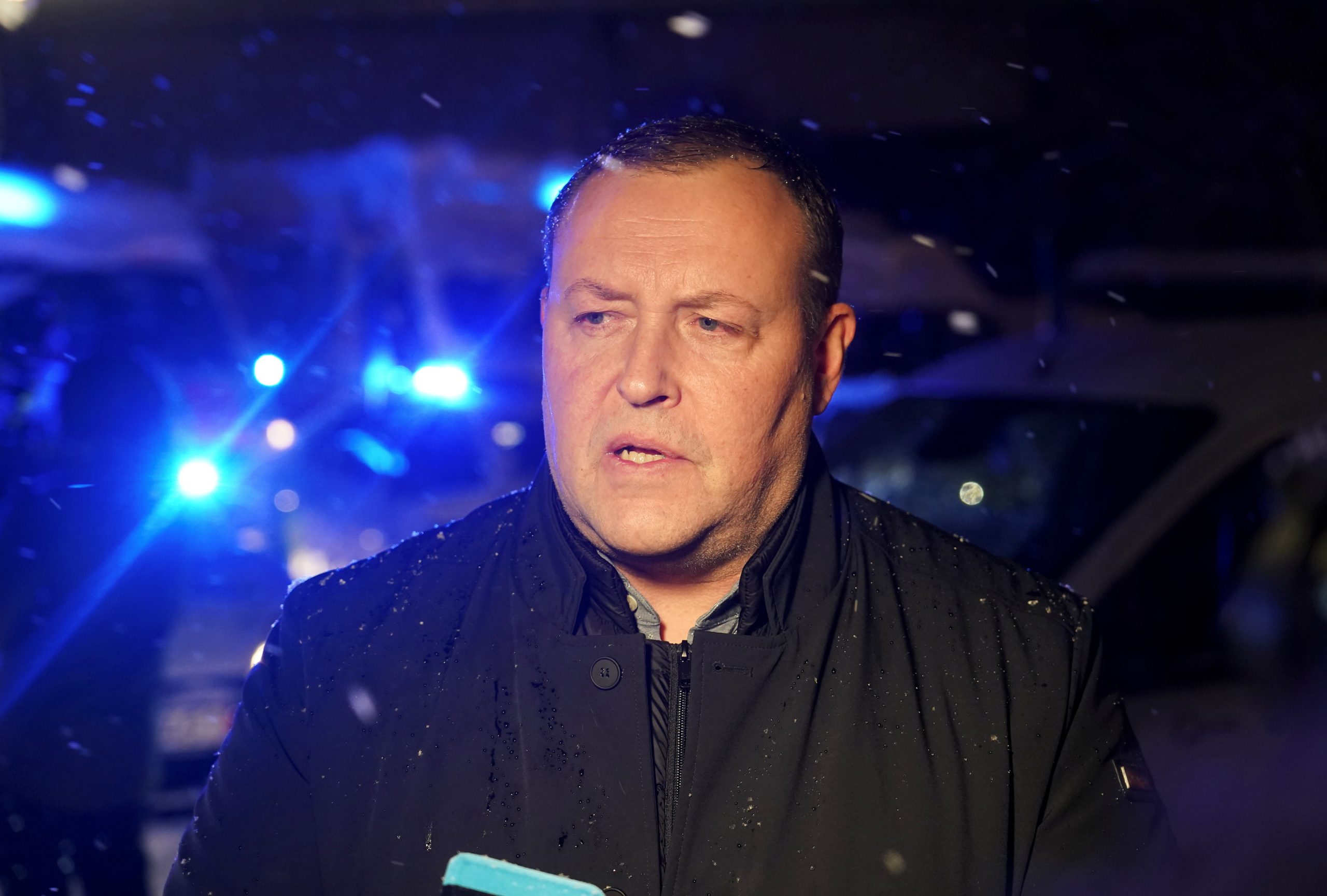 Iekšlietu ministrs Rihards Kozlovskis melnā jakā sniedz interviju vēlā vakara stundā, fonā redzami mirgojoši policijas gaismas signāli un krītošs sniegs