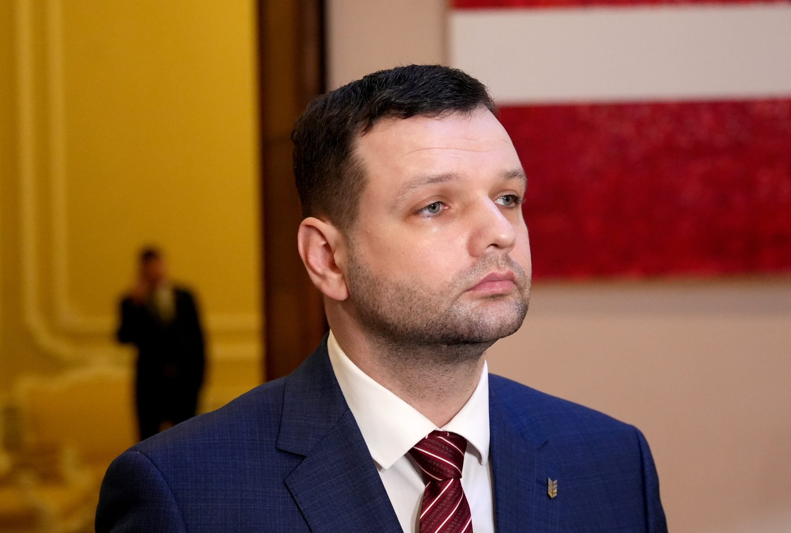 Labklājības ministrs Reinis Uzulnieks tumši zilā uzvalkā, fonā Latvijas karogs