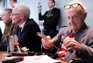 Vecāka gadagājuma vīrietis ar brillēm, rokās turot pašdarinātu priekšmetu, sēžot pie galda publiskā pasākumā