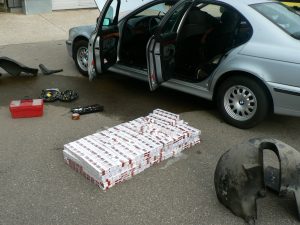 Sudraba krāsas automašīna ar atvērtām durvīm un noņemtiem detaļu paneļiem, blakus uz zemes redzams liels cigarešu bloku klāsts