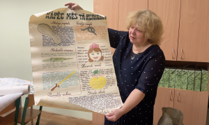 Sieviete tur rokās lielu, ar roku rakstītu un ilustrētu plakātu par izteicieniem latviešu valodā