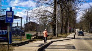Sieviete gaišā jakā iet gar autobusa pieturu ar uzrakstu Kultūras nams, apkārt pavasarīgas ielas ainava