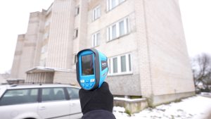 Rokā turēta termokamera, vērsta pret daudzdzīvokļu mājas fasādi ziemā.