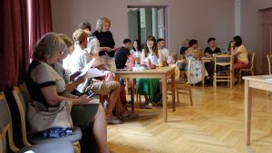 Dažāda vecuma cilvēki piedalās diskusijā vai apmācībās, sēžot pie galdiem kopienas telpā.