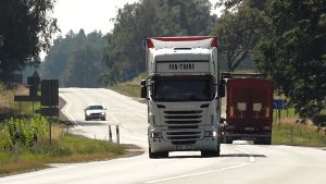 Kravas automašīnas un vieglais auto brauc pa lauku šoseju saulainā dienā.