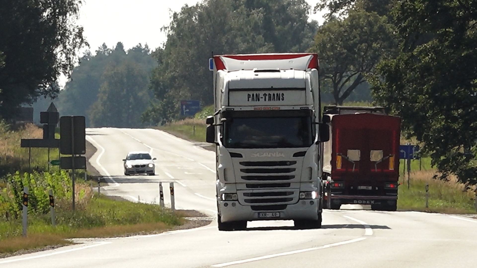 Kravas automašīnas un vieglais auto brauc pa lauku šoseju saulainā dienā.