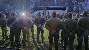 Kadeti militārā formā stāv ierindā ārā, kamēr instruktors uzrunā viņus pie autobusa.