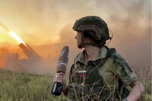 Sieviete militārā formā ar Krievijas karoga uzšuvi un preses mikrofonu, kamēr fonā redzams raķešu palaišanas uzliesmojums lauka apstākļos