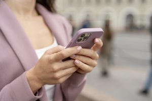 Sieviete rozā žaketē tur viedtālruni rozā maciņā uz pilsētas fona