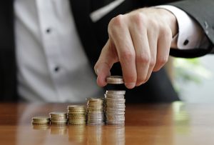 Biznesmenis skaita monētu kaudzes, simbolizējot finanšu izaugsmi un uzkrājumus.