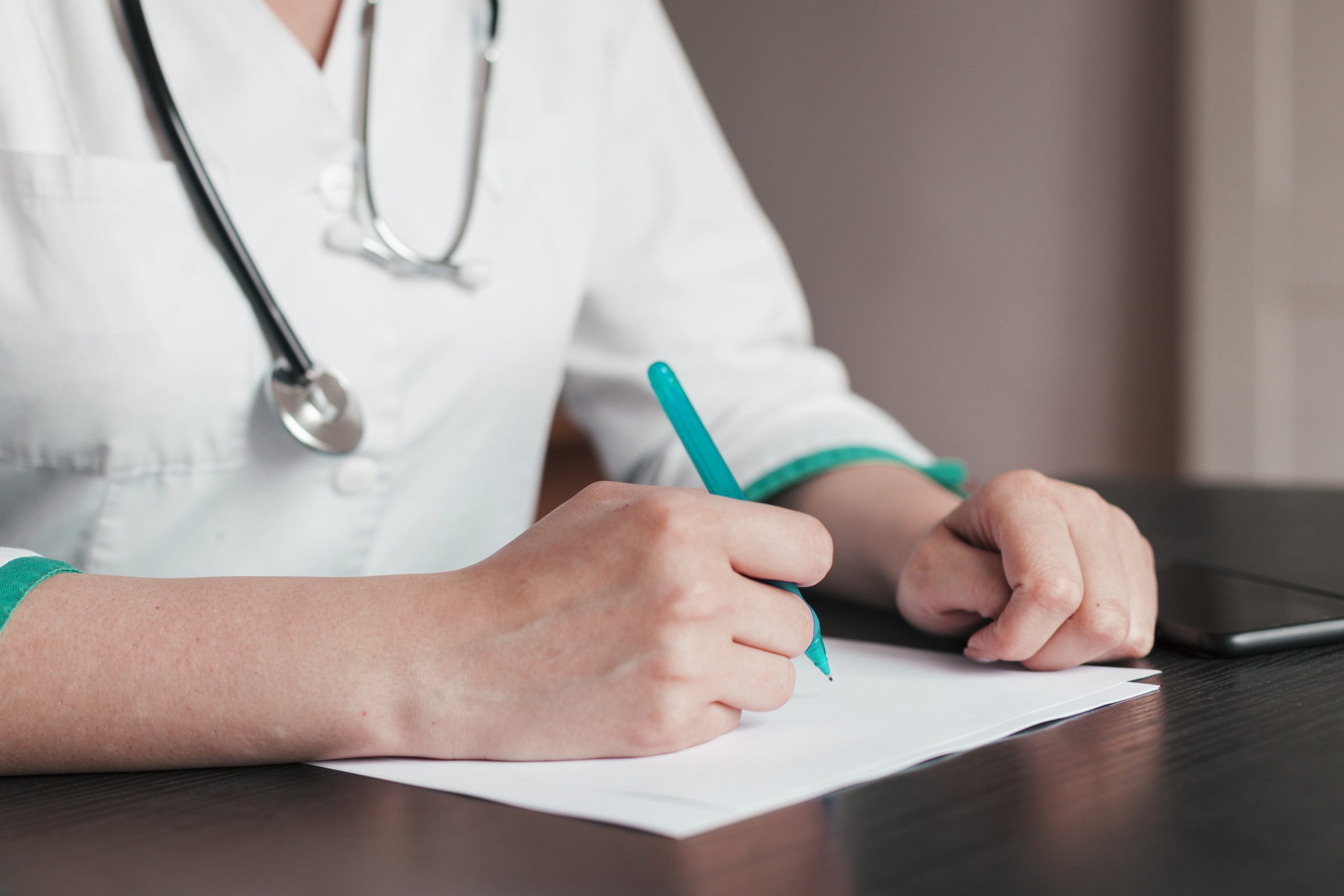 Ārsts baltā halātā ar stetoskopu raksta medicīnisku ziņojumu.
