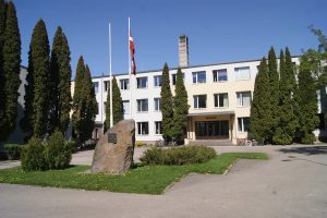 Gaiša trīs stāvu ēka ar baltām un bēšām fasādes daļām, priekšpusē liels piemiņas akmens un Latvijas karogs, apkārt aug gari dekoratīvie koki.