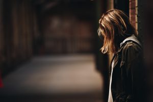 Skumja jauna sieviete ar nolaistu galvu, atspiedusies pret sienu tumšā ejā.