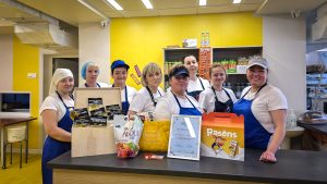 Vairākas ēdnīcas darbinieces baltos krekliņos un zilās priekšautos, stāvot pie letes un prezentējot dažādus pārtikas produktus