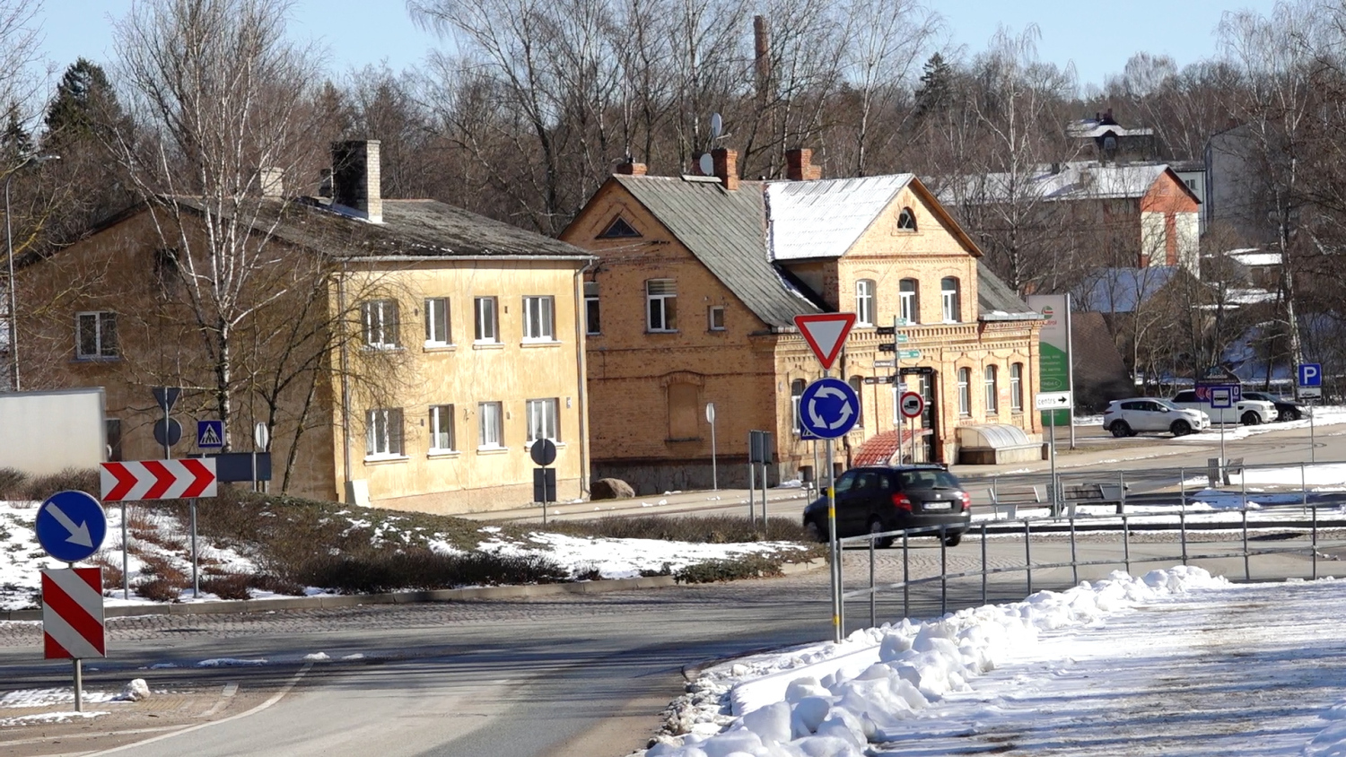 Vairākas dzeltenas un ķieģeļu ēkas pie sniegota, apļveida krustojuma ar ceļa zīmēm un braucošu automašīnu