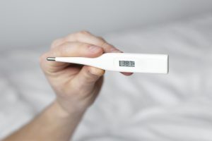 Rokā turēts digitālais termometrs ar temperatūras rādījumu 38,7°C.