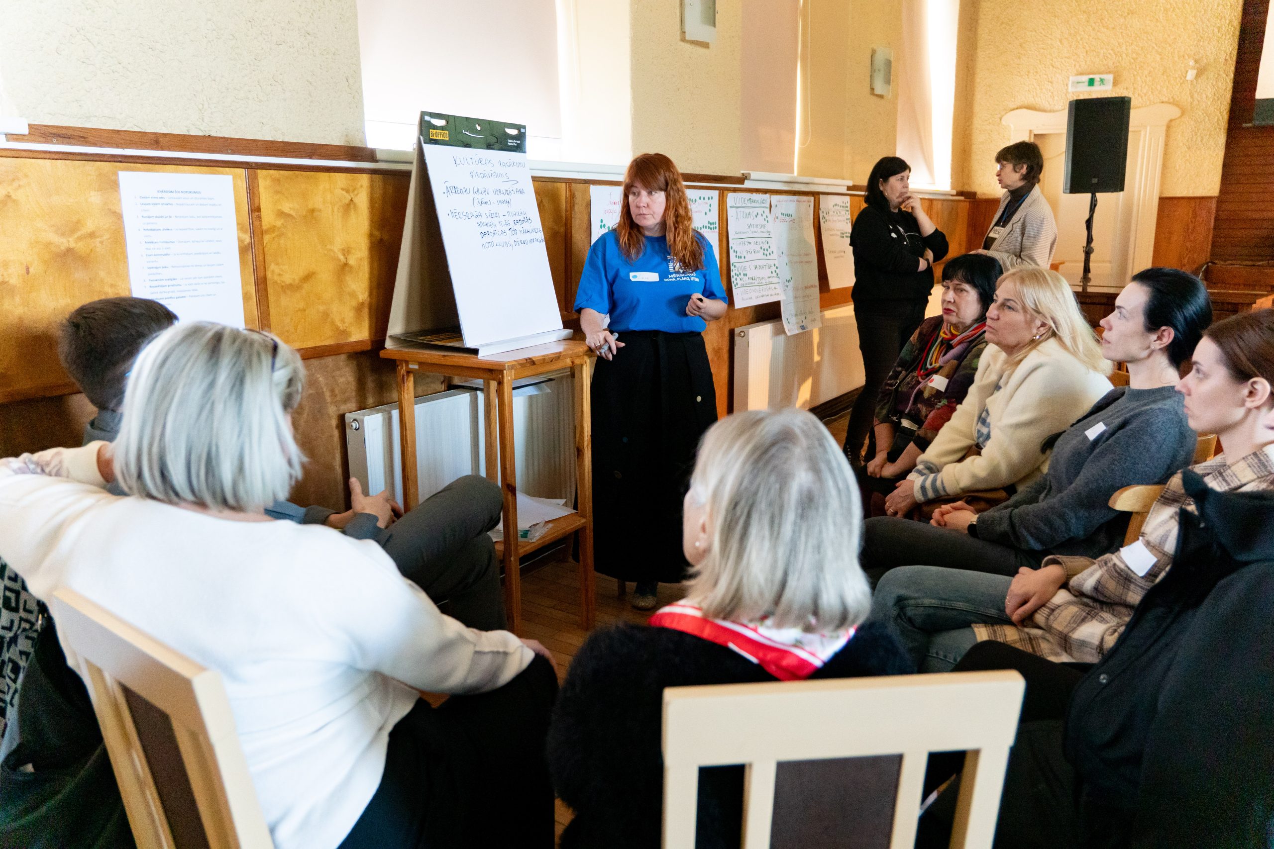 Grupas diskusija seminārā – sieviete zilā kreklā vada prezentāciju, kamēr dalībnieki klausās