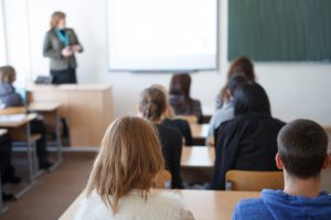 Studenti auditorijā klausās lekciju, pasniedzēja priekšā pie tāfeles.