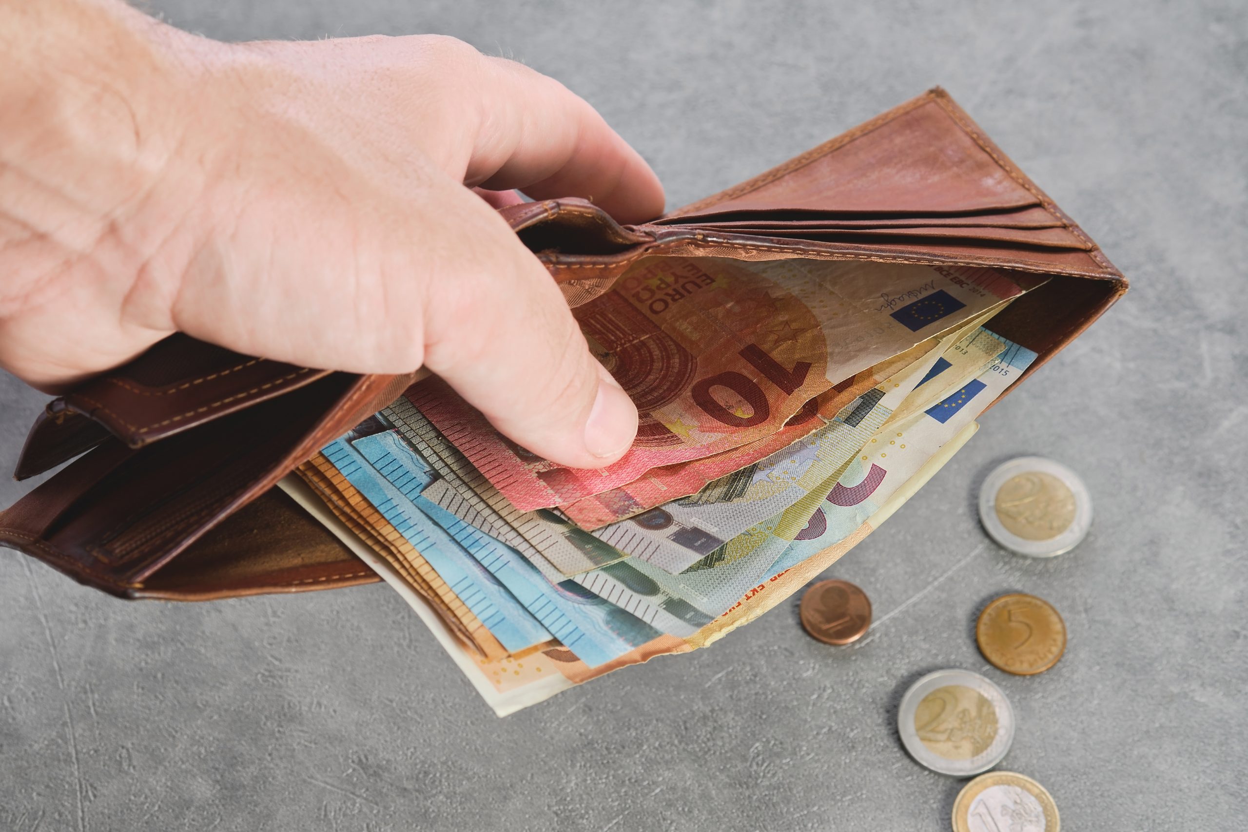 Rokā turēts brūns maks, pilns ar eiro banknotēm, ar monētām uz galda fonā