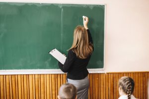 Skolotāja raksta uz tāfeles klasē, priekšplānā skolēni.